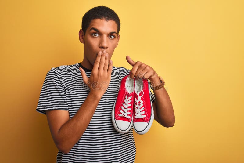 Junge, gut aussehende Arab-Mann, die lockere Sneakers über einem isolierten, gelben Hintergrund halten, mit der Hand schockiert mi lizenzfreie stockfotos