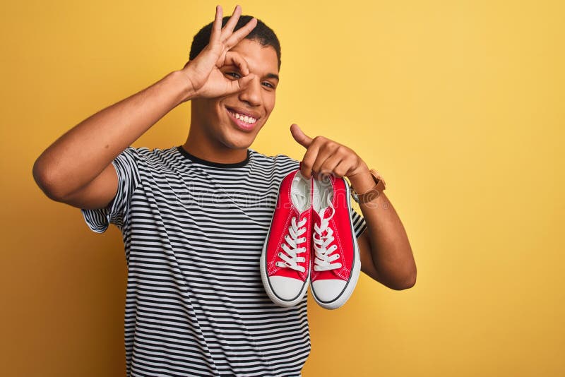 Junge, gut aussehende Arab-Mann, die lockere Sneakers über isoliertem gelbem Hintergrund hält, mit einem glücklichen Gesicht, der lizenzfreie stockfotografie