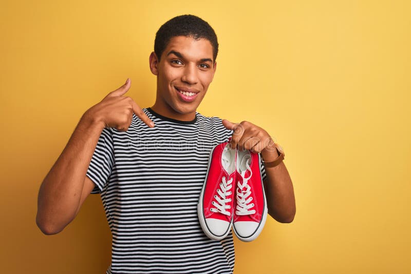 Junge, gut aussehende Arab-Mann, die lockere Sneakers über isoliertem gelbem Hintergrund hält, mit einem überraschenden Zeigefinge stockfoto