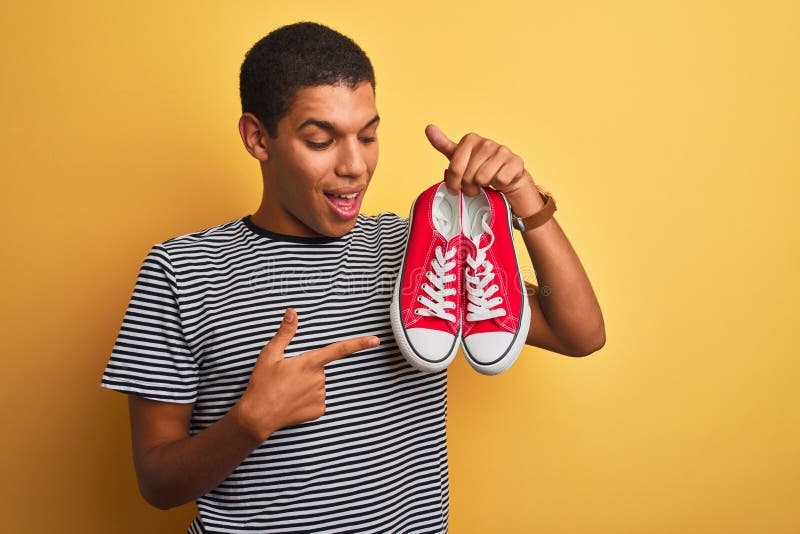 Junge, gut aussehende Arab-Mann, die lockere Sneakers über isoliertem gelbem Hintergrund hält und mit Hand und Finger sehr glückli stockbilder