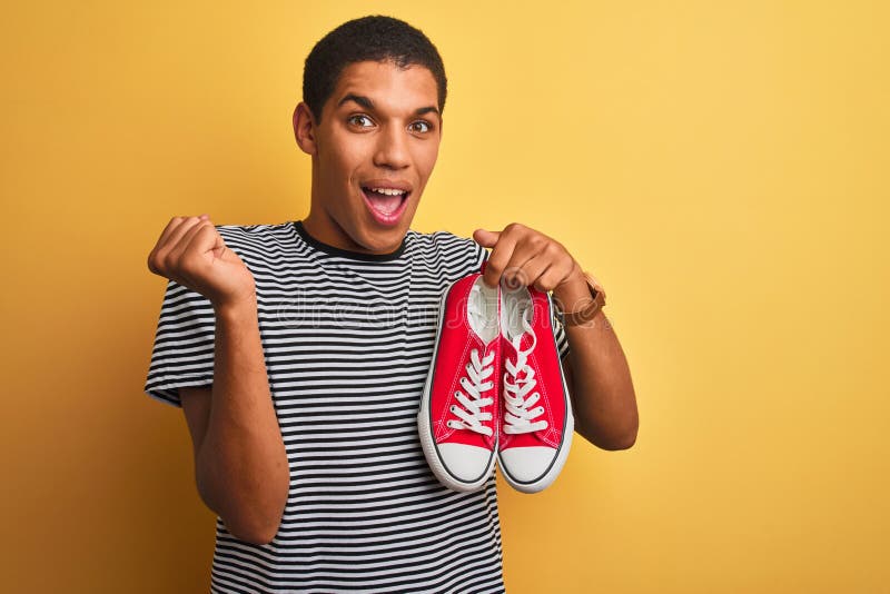 Junge, gut aussehende Araber, die über einem isolierten gelben Hintergrund lockere Sneakers halten und stolz schreien und den Sieg lizenzfreie stockbilder