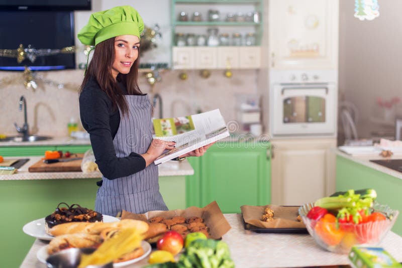 Junge Hausfrau, die versucht, ein neues Rezept im Kochbuch bei mit Lebensmittel und Bestandteilen bei Tisch stehen zu finden lizenzfreies stockfoto Junge Hausfrau, die versucht, ein neues Rezept im Kochbuch bei mit Lebensmittel und Bestandteilen bei Tisch stehen zu finden lizenzfreies stockfoto