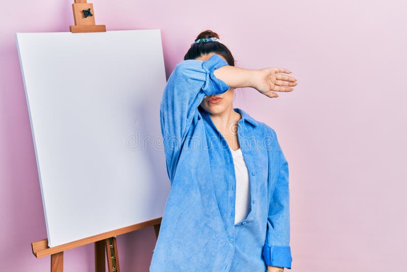 Young hispanic woman standing by painter easel stand covering eyes with arm, looking serious and sad. sightless, hiding and rejection concept. Young hispanic woman standing by painter easel stand covering eyes with arm, looking serious and sad. sightless, hiding and rejection concept