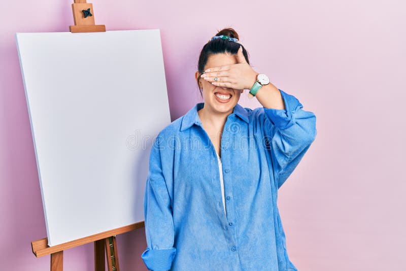 Young hispanic woman standing by painter easel stand smiling and laughing with hand on face covering eyes for surprise. blind concept. Young hispanic woman standing by painter easel stand smiling and laughing with hand on face covering eyes for surprise. blind concept
