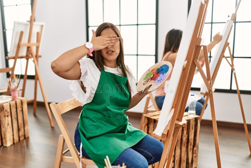 Young hispanic artist women painting on canvas at art studio covering eyes with hand, looking serious and sad. sightless, hiding and rejection concept. Young hispanic artist women painting on canvas at art studio covering eyes with hand, looking serious and sad. sightless, hiding and rejection concept