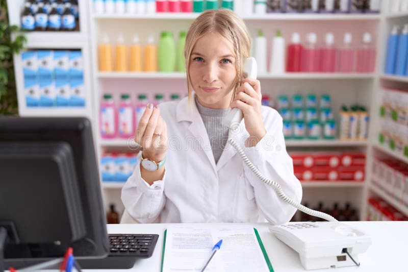 Young caucasian woman working at pharmacy drugstore speaking on the telephone doing money gesture with hands, asking for salary payment, millionaire business. Young caucasian woman working at pharmacy drugstore speaking on the telephone doing money gesture with hands, asking for salary payment, millionaire business