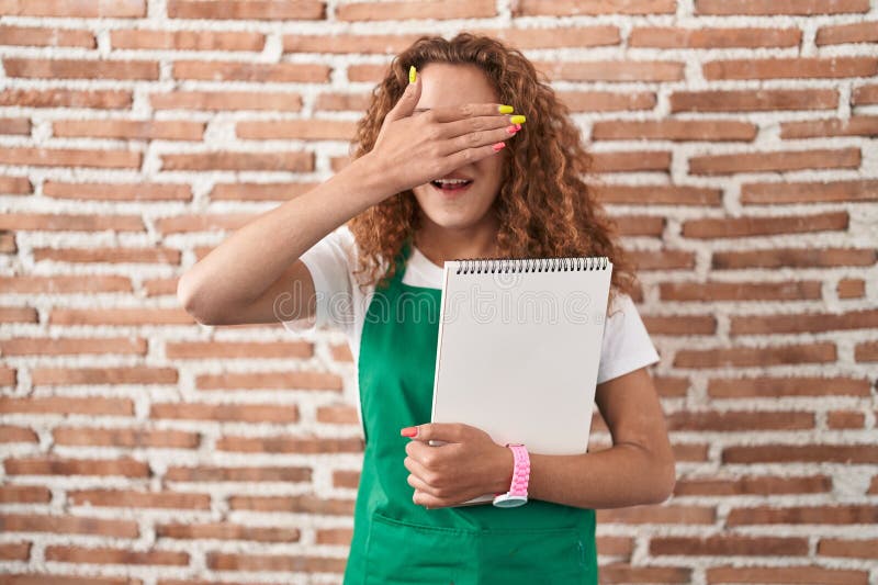 Young caucasian woman holding art notebook smiling and laughing with hand on face covering eyes for surprise. blind concept. Young caucasian woman holding art notebook smiling and laughing with hand on face covering eyes for surprise. blind concept