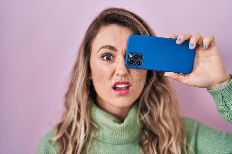 Young caucasian woman covering eye with smartphone clueless and confused expression. doubt concept. Young caucasian woman covering eye with smartphone clueless and confused expression. doubt concept