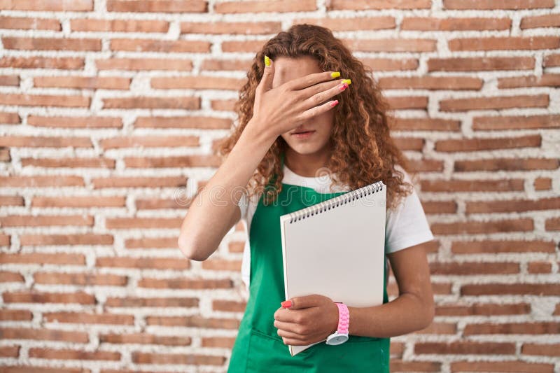 Young caucasian woman holding art notebook covering eyes with hand, looking serious and sad. sightless, hiding and rejection concept. Young caucasian woman holding art notebook covering eyes with hand, looking serious and sad. sightless, hiding and rejection concept