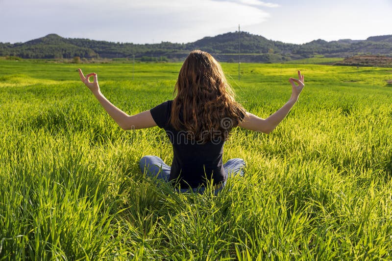 Young caucasian woman red-haired with freckles, resting on a green field at spring sunset, sitting in a yoga position. Meditation, mindfulness, relax. Young caucasian woman red-haired with freckles, resting on a green field at spring sunset, sitting in a yoga position. Meditation, mindfulness, relax