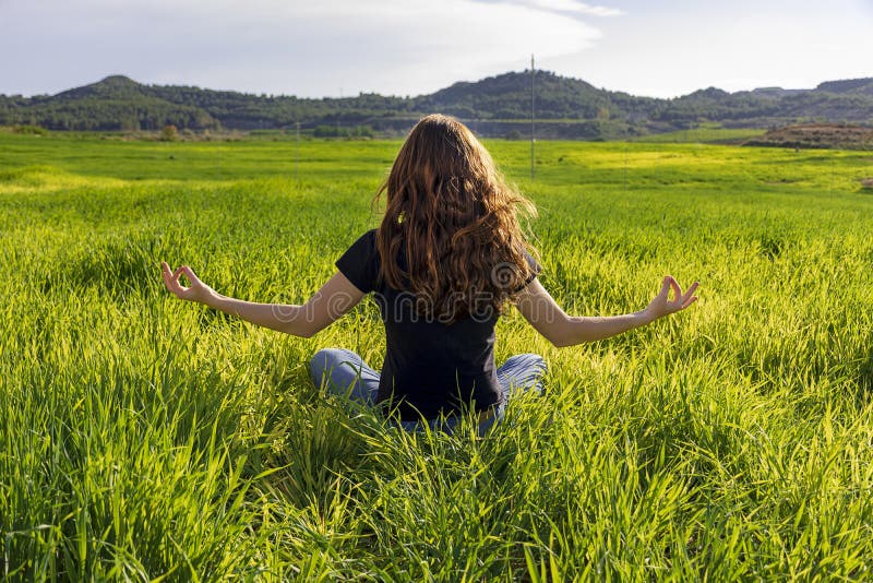 Young caucasian woman red-haired with freckles, resting on a green field at spring sunset, sitting in a yoga position. Meditation, mindfulness, relax. Young caucasian woman red-haired with freckles, resting on a green field at spring sunset, sitting in a yoga position. Meditation, mindfulness, relax