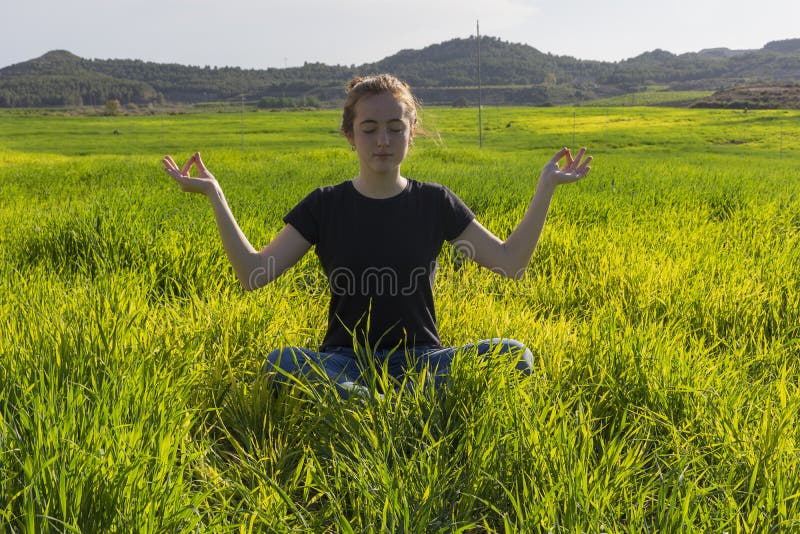 Young caucasian woman red-haired with freckles, resting on a green field at spring sunset, sitting in a yoga position. Meditation, mindfulness, relax. Young caucasian woman red-haired with freckles, resting on a green field at spring sunset, sitting in a yoga position. Meditation, mindfulness, relax