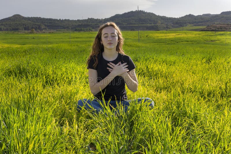 Young caucasian woman red-haired with freckles, resting on a green field at spring sunset, sitting in a yoga position. Meditation, mindfulness, relax. Young caucasian woman red-haired with freckles, resting on a green field at spring sunset, sitting in a yoga position. Meditation, mindfulness, relax