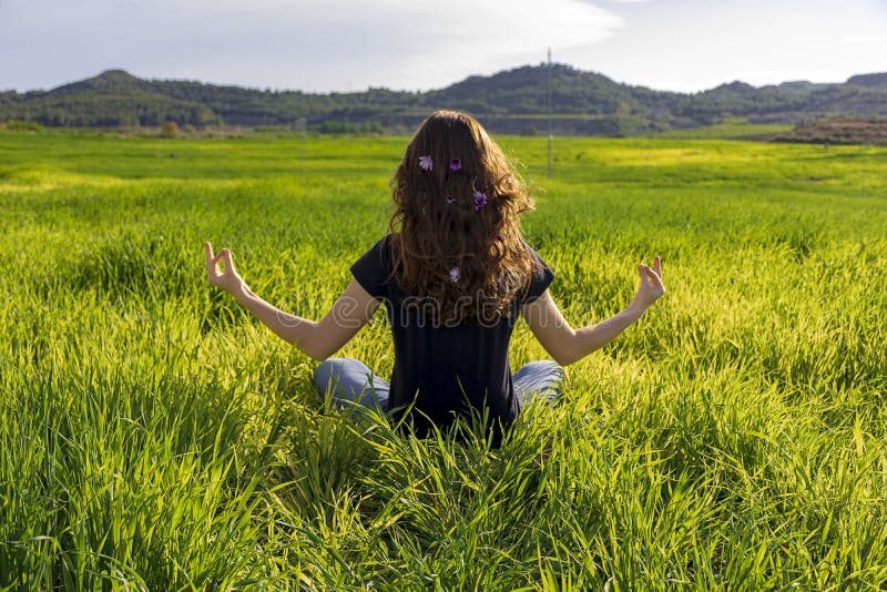 Young caucasian woman red-haired with freckles, resting on a green field at spring sunset, sitting in a yoga position. Meditation, mindfulness, relax. Young caucasian woman red-haired with freckles, resting on a green field at spring sunset, sitting in a yoga position. Meditation, mindfulness, relax