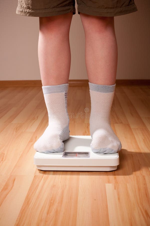 Boy measures weight on floor scales. Legs in shorts and socks standing at floor scales on hardwood floor in living room. Boy measures weight on floor scales. Legs in shorts and socks standing at floor scales on hardwood floor in living room.