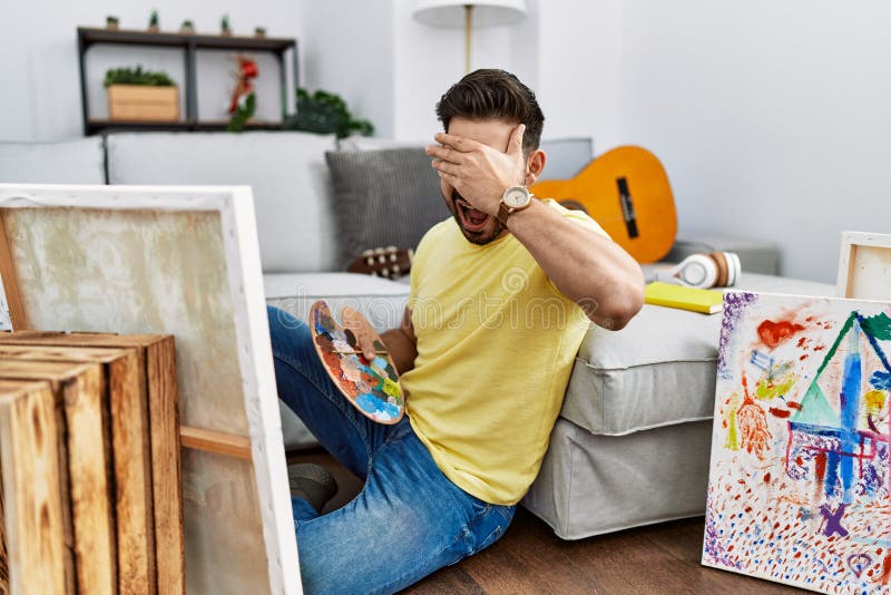 Young man with beard painting canvas at home smiling and laughing with hand on face covering eyes for surprise. blind concept. Young man with beard painting canvas at home smiling and laughing with hand on face covering eyes for surprise. blind concept