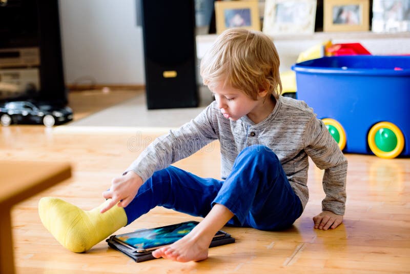 Cute little blond boy with broken leg in cast sitting on the wooden floor, playing on tablet. Child's daytime fun. Happy to be at home. Cute little blond boy with broken leg in cast sitting on the wooden floor, playing on tablet. Child's daytime fun. Happy to be at home.