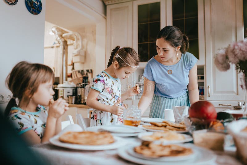 Junge mitfühlende Mutter und ihre zwei kleine Töchter, die Pfannkuchen mit Honig am Frühstück in der gemütlichen Küche essen lizenzfreie stockfotografie Junge mitfühlende Mutter und ihre zwei kleine Töchter, die Pfannkuchen mit Honig am Frühstück in der gemütlichen Küche essen lizenzfreie stockfotografie