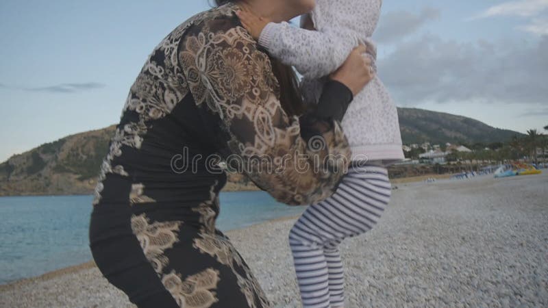 Junge Mutter mit Baby auf dem Sonnenuntergangstrand Attraktive kaukasische Frau im schwarzen festen Kleid dehnt Handdas anheben a
