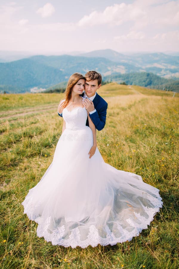 Junge romantische Heiratspaare, die auf sonniger Rasenfläche mit entferntem Forest Hills und erstaunlichem Himmel als Hintergrund lizenzfreie stockbilder Junge romantische Heiratspaare, die auf sonniger Rasenfläche mit entferntem Forest Hills und erstaunlichem Himmel als Hintergrund lizenzfreie stockbilder