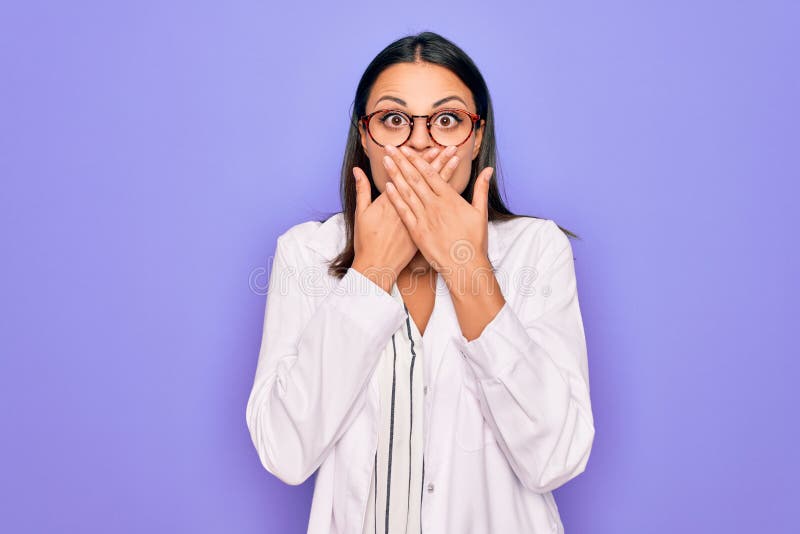 Junge schöne Brunette-Psychologin mit Mantel und Brille über violettem Hintergrund schockiert mit Händen für lizenzfreie stockbilder