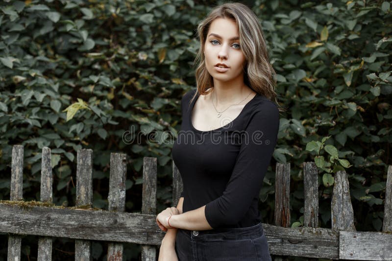 Junge schöne hübsche nette ländliche Frau in den modernen Jeans in einem stilvollen schwarzen T-Shirt steht nahe einem hölzernen stockfoto Junge schöne hübsche nette ländliche Frau in den modernen Jeans in einem stilvollen schwarzen T-Shirt steht nahe einem hölzernen stockfoto