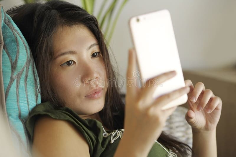 Junge schöne und entspannte asiatische Chinesin, die zu Hause Wohnzimmersofacouch unter Verwendung des Internets auf dem Handy gl lizenzfreie stockfotografie Junge schöne und entspannte asiatische Chinesin, die zu Hause Wohnzimmersofacouch unter Verwendung des Internets auf dem Handy gl lizenzfreie stockfotografie