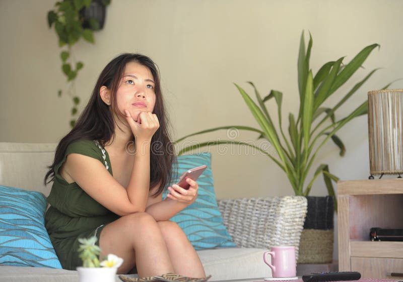 Junge schöne und entspannte asiatische Chinesin, die zu Hause Wohnzimmersofacouch unter Verwendung des Internets auf dem Handy gl lizenzfreies stockbild Junge schöne und entspannte asiatische Chinesin, die zu Hause Wohnzimmersofacouch unter Verwendung des Internets auf dem Handy gl lizenzfreies stockbild