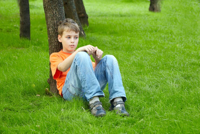 Boy in orange t-shirt sits with thoughtful face on grass, leaning his back on trunk of tree. Boy in orange t-shirt sits with thoughtful face on grass, leaning his back on trunk of tree