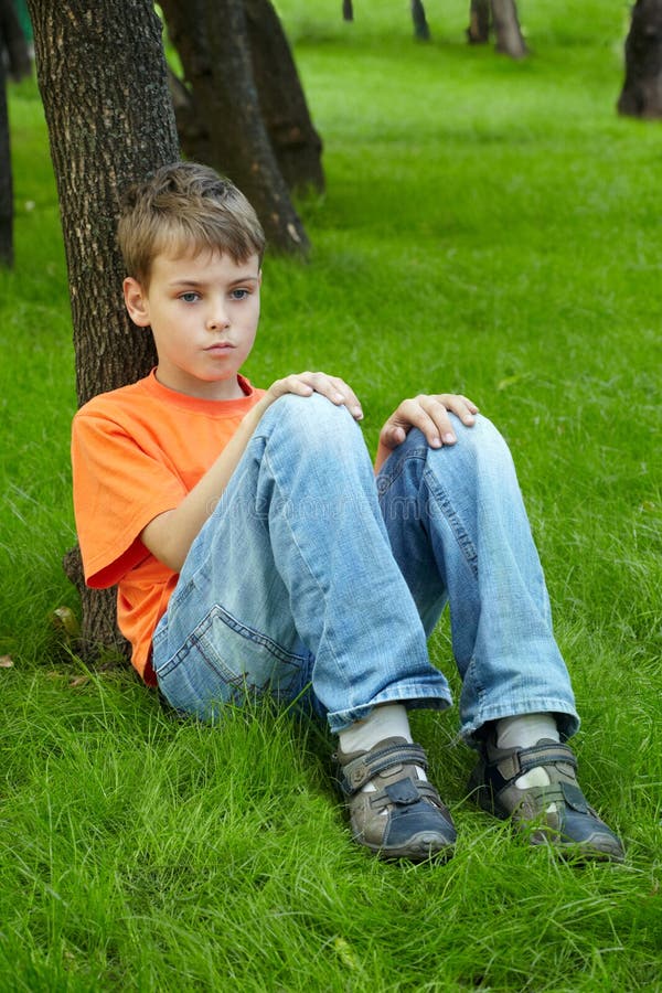 Boy in orange t-shirt sits with thoughtful face on grass, looking in front of himself and leaning his back on trunk of tree. Boy in orange t-shirt sits with thoughtful face on grass, looking in front of himself and leaning his back on trunk of tree