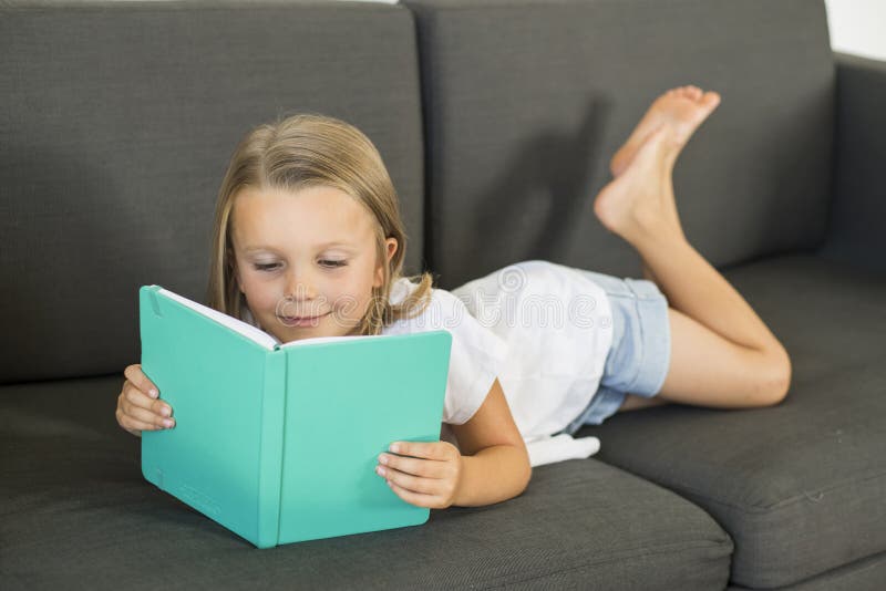 Junge süße und glückliche kleine alte Lügen des Mädchens 6 oder 7 Jahre auf der Hauptwohnzimmersofacouch, die eine Buchruhe und e lizenzfreies stockbild Junge süße und glückliche kleine alte Lügen des Mädchens 6 oder 7 Jahre auf der Hauptwohnzimmersofacouch, die eine Buchruhe und e lizenzfreies stockbild