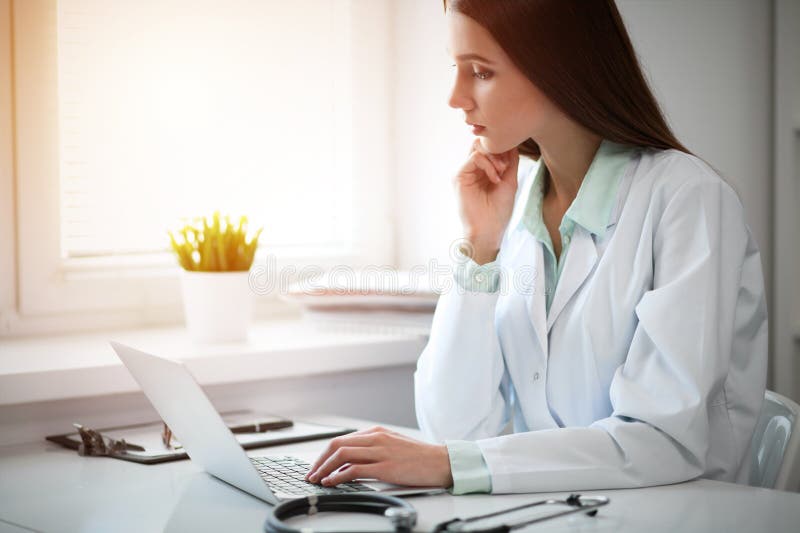 Junge Ärztin, die auf Laptop-Computer beim Sitzen am Tisch nahe dem Fenster im Krankenhausbüro schreibt lizenzfreie stockfotografie Junge Ärztin, die auf Laptop-Computer beim Sitzen am Tisch nahe dem Fenster im Krankenhausbüro schreibt lizenzfreie stockfotografie