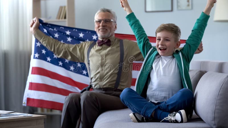 Jungen-und Großvater wellenartig bewegende amerikanische Flagge, aufpassendes Sportspiel zu Hause, glücklich für Gewinn lizenzfreie stockfotografie Jungen-und Großvater wellenartig bewegende amerikanische Flagge, aufpassendes Sportspiel zu Hause, glücklich für Gewinn lizenzfreie stockfotografie