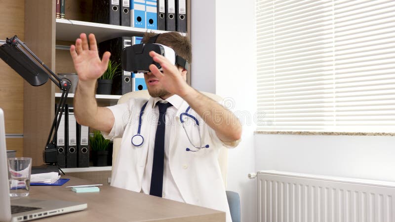 Junger Arzt in der weißen Robe und in der blauen Bindung in seinem Büro trägt einen Kopfhörer der virtuellen Realität VR stock video footage