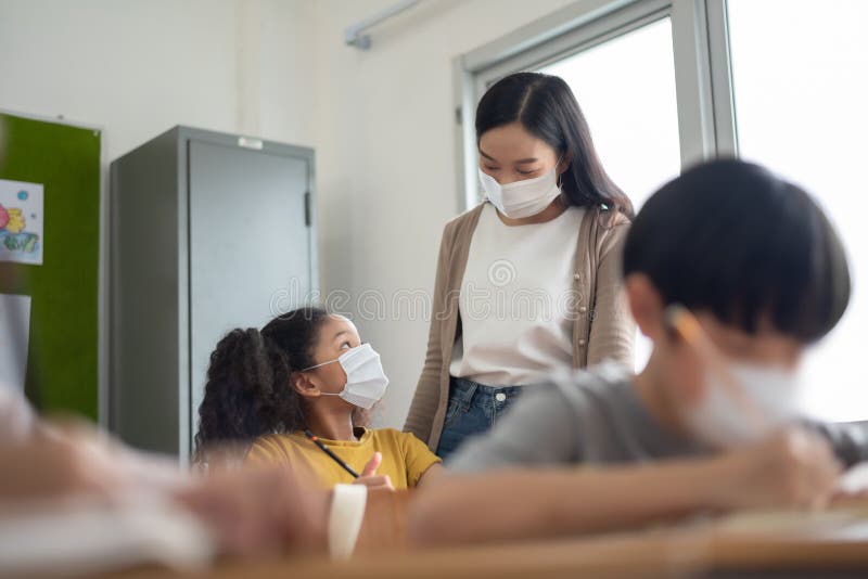Junger asiatischer Lehrer mit dem africanamerican Mädchen, das den Schutz der Gesichtsmasken Prüfung im Klassenzimmer trägt lizenzfreies stockbild