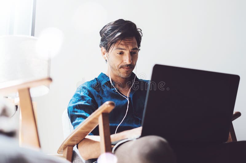 Junger attraktiver Mann, der im sonnigen Büro auf Laptop arbeitet Bemannen Sie der Notizbuchtastatur an zeigen und Musik hören stockfotografie Junger attraktiver Mann, der im sonnigen Büro auf Laptop arbeitet Bemannen Sie der Notizbuchtastatur an zeigen und Musik hören stockfotografie