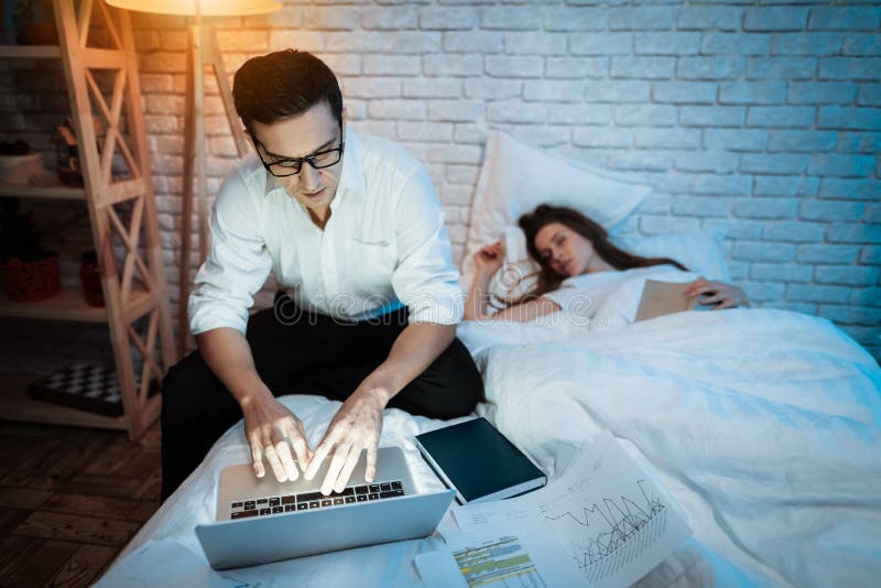 Junger Geschäftsmann, der an Laptop im Bett mit junger Frau arbeitet Junge Frau schläft stockfotografie Junger Geschäftsmann, der an Laptop im Bett mit junger Frau arbeitet Junge Frau schläft stockfotografie