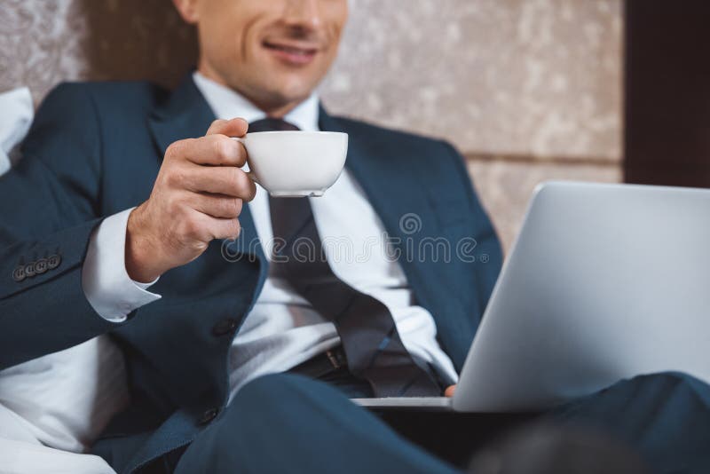 Junger Geschäftsmann im Gesellschaftsanzug unter Verwendung seines Laptops und halten Tasse Kaffees, beim Sitzen auf Bett stockfotografie Junger Geschäftsmann im Gesellschaftsanzug unter Verwendung seines Laptops und halten Tasse Kaffees, beim Sitzen auf Bett stockfotografie