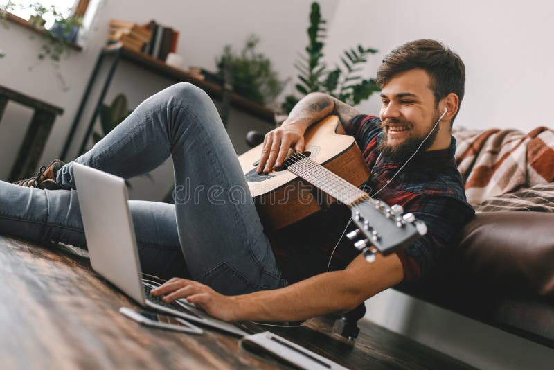 Junger Gitarristhippie zu Hause, der auf dem Boden hält Gitarrenkopfhörermusikgrasenlaptop sitzt stockfoto Junger Gitarristhippie zu Hause, der auf dem Boden hält Gitarrenkopfhörermusikgrasenlaptop sitzt stockfoto
