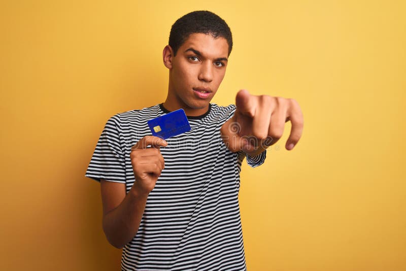 Junger, gut aussehender arabischer Mann mit Kreditkarte über isoliertem gelbem Hintergrund, der mit dem Finger auf die Kamera und stockfotografie