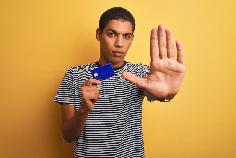 Junger, gut aussehender arabischer Mann mit Kreditkarte über isoliertem gelbem Hintergrund mit offener Hand, Stoppschild mit ernst lizenzfreie stockfotos