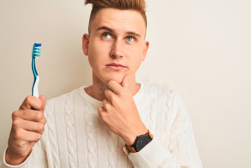Junger gut aussehender Mann, der Zahnbürste über isoliertem weißen Hintergrund hält, ernstes Gesicht, das über Fragen nachdenkt, s lizenzfreies stockfoto