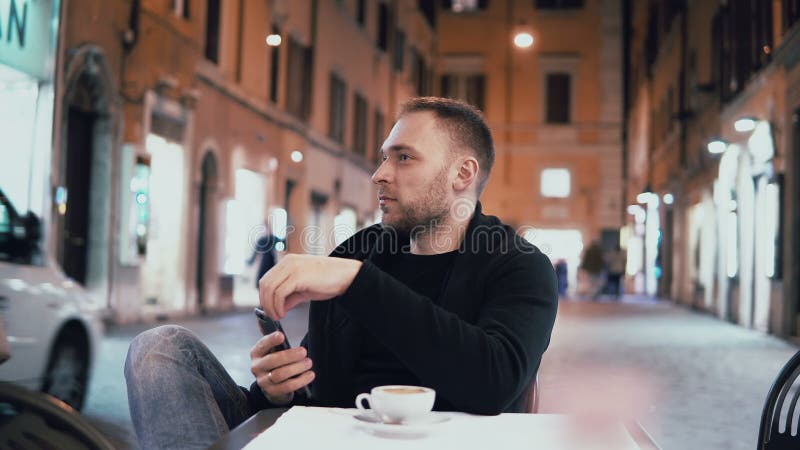Junger gutaussehender Mann, der draußen im Stadtcafé sitzt und Smartphone hält Mannestrinkender Kaffee am Abend stockfotografie Junger gutaussehender Mann, der draußen im Stadtcafé sitzt und Smartphone hält Mannestrinkender Kaffee am Abend stockfotografie