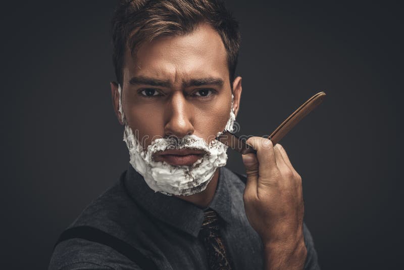Junger gutaussehender Mann mit Rasierschaum auf seinem Gesicht, seinen Bart mit geradem Rasiermesser und dem Schauen pflegend lizenzfreie stockfotografie