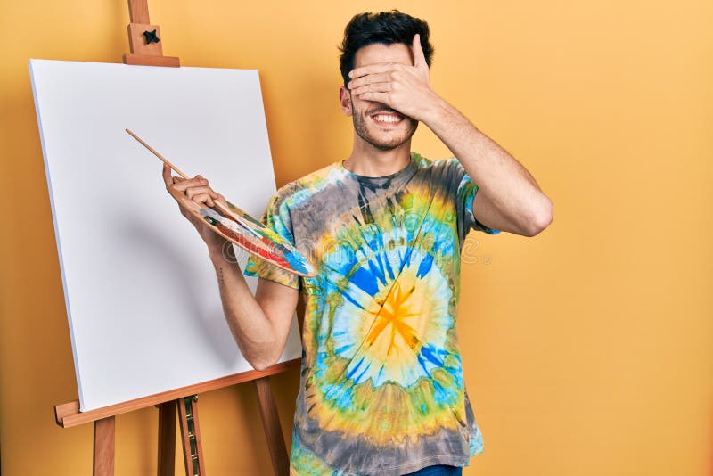 Young hispanic man standing drawing with palette by painter easel stand smiling and laughing with hand on face covering eyes for surprise. blind concept. Young hispanic man standing drawing with palette by painter easel stand smiling and laughing with hand on face covering eyes for surprise. blind concept