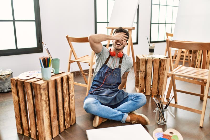 Young hispanic man sitting at art studio smiling and laughing with hand on face covering eyes for surprise. blind concept. Young hispanic man sitting at art studio smiling and laughing with hand on face covering eyes for surprise. blind concept