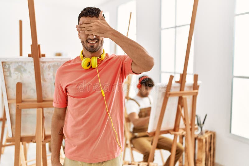 Young hispanic man at art studio smiling and laughing with hand on face covering eyes for surprise. blind concept. Young hispanic man at art studio smiling and laughing with hand on face covering eyes for surprise. blind concept