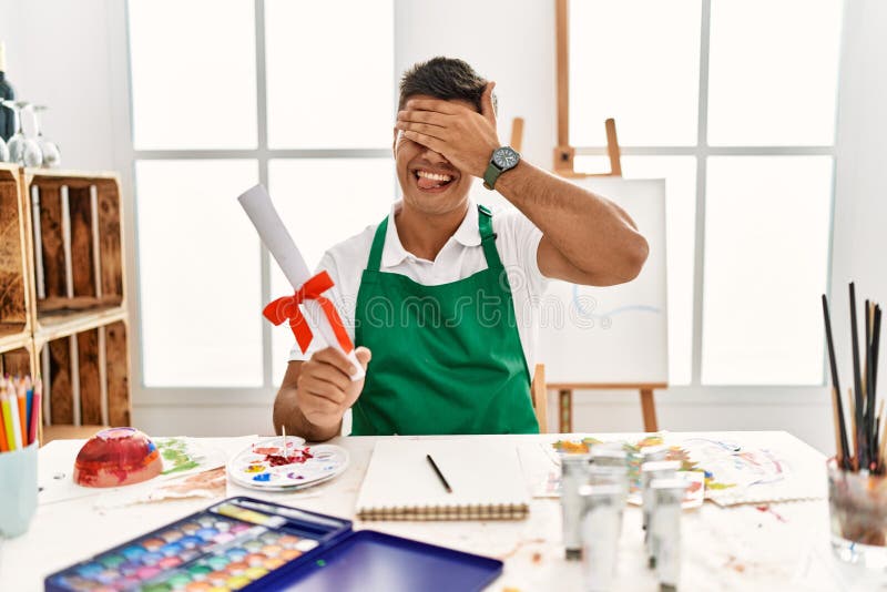 Young hispanic man at art studio holding degree smiling and laughing with hand on face covering eyes for surprise. blind concept. Young hispanic man at art studio holding degree smiling and laughing with hand on face covering eyes for surprise. blind concept