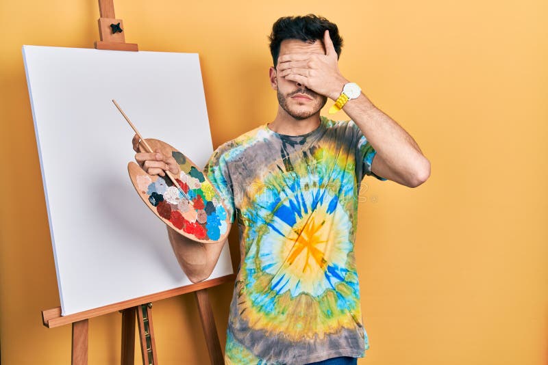 Young hispanic man standing drawing with palette by painter easel stand covering eyes with hand, looking serious and sad. sightless, hiding and rejection concept. Young hispanic man standing drawing with palette by painter easel stand covering eyes with hand, looking serious and sad. sightless, hiding and rejection concept