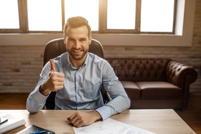 Junger hübscher netter Geschäftsmann sitzen auf Tabelle und werfen in seinem eigenen Büro auf Er hält großen Daumen und Lächeln h lizenzfreie stockfotografie Junger hübscher netter Geschäftsmann sitzen auf Tabelle und werfen in seinem eigenen Büro auf Er hält großen Daumen und Lächeln h lizenzfreie stockfotografie